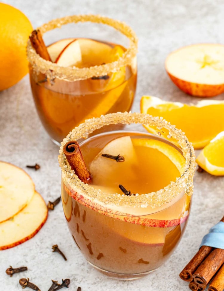 two glasses with homemade apple cider garnished with a cinnamon stick and a few apple slices.