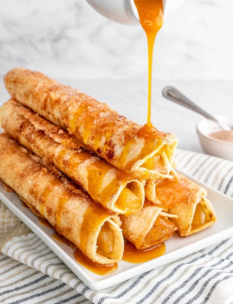 a stack of apple pie taquitos on a white plate drizzled with caramel sauce.