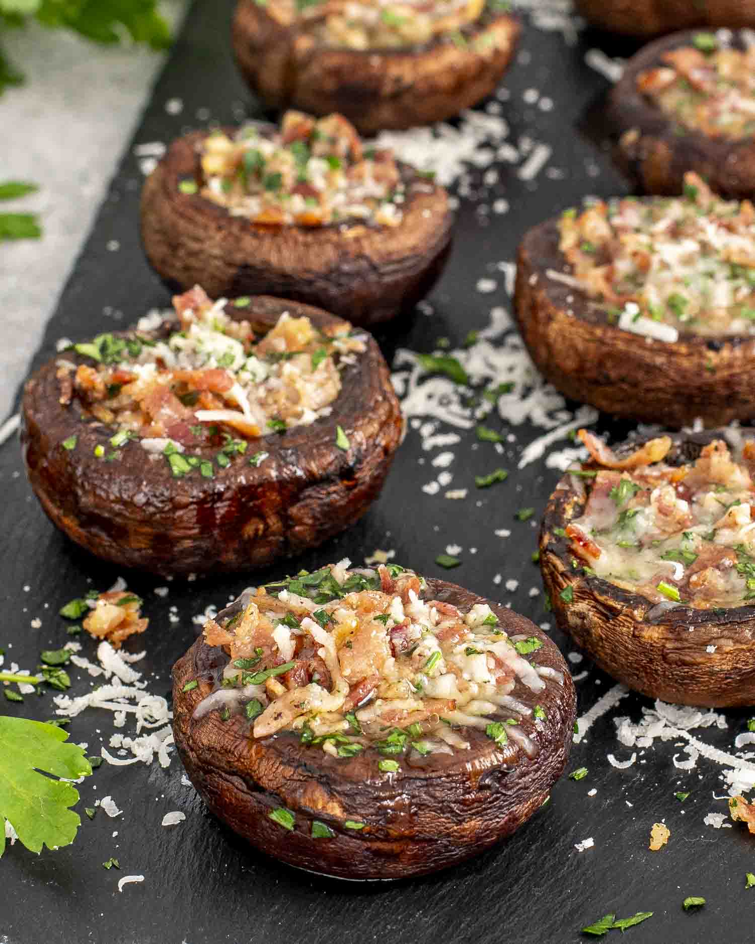 Close-up of Bacon Stuffed Mushrooms arranged on a dark serving platter. The cremini mushrooms are filled with a savory mixture of crispy bacon, melted Parmesan cheese, and fresh parsley. 