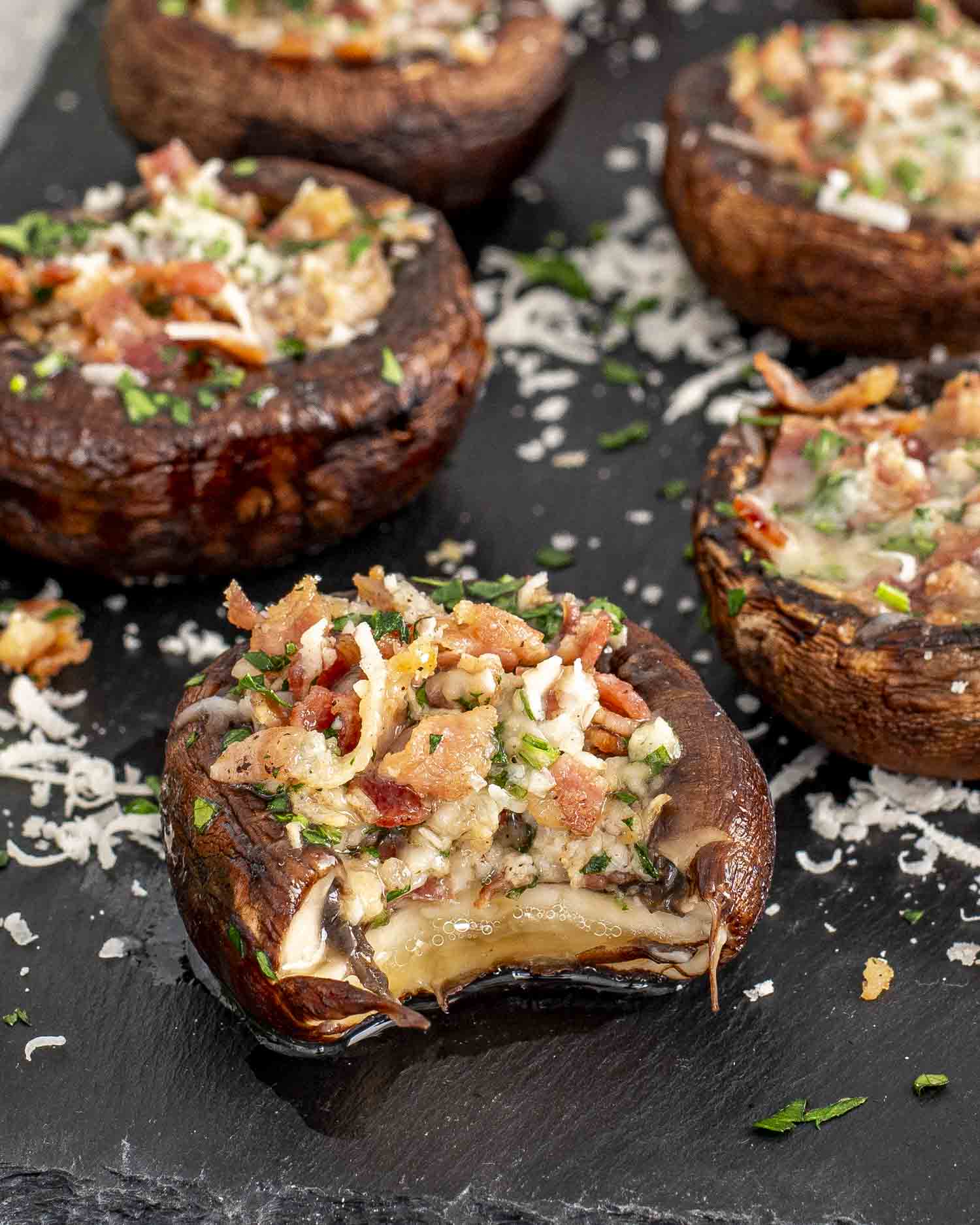 Close-up of Bacon Stuffed Mushrooms arranged on a dark serving platter. The cremini mushrooms are filled with a savory mixture of crispy bacon, melted Parmesan cheese, and fresh parsley.