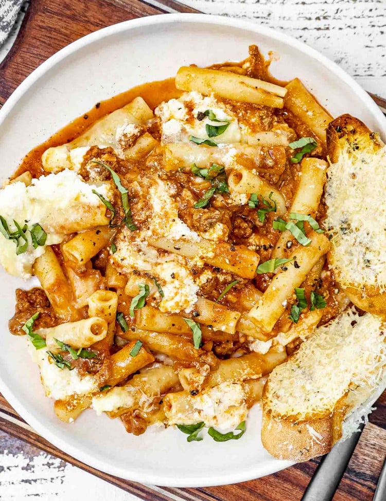 a big bowl of baked ziti garnished with basil and a couple slices of toasted bread.