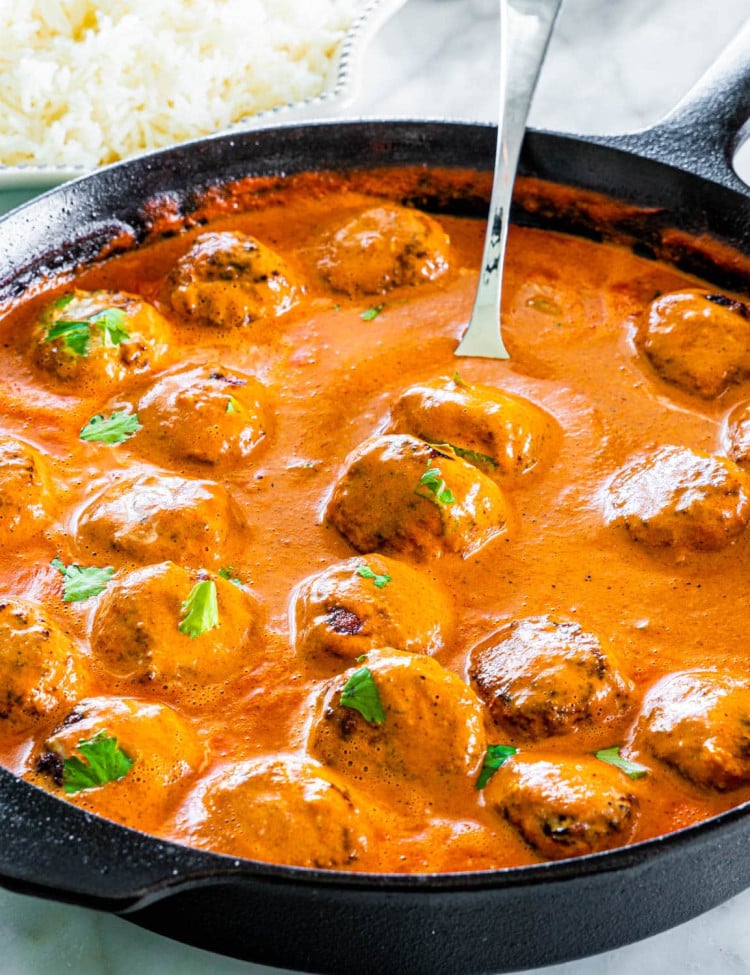 side shot of butter chicken meatballs in a black skillet with a serving spoon inside