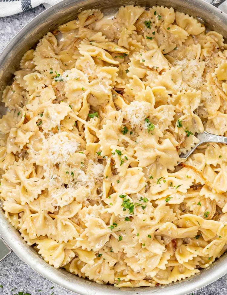 A close-up image of a skillet filled with bowtie pasta tossed in a creamy, caramelized onion sauce, garnished with fresh parsley and grated Parmesan cheese.