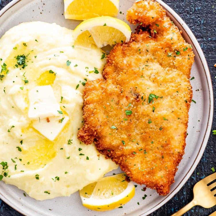 a chicken schnitzel on a plate with mashed potatoes and lemon wedges.