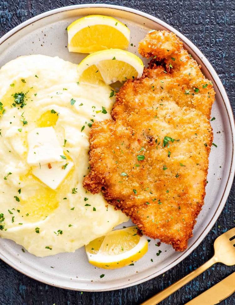 a chicken schnitzel on a plate with mashed potatoes and lemon wedges.