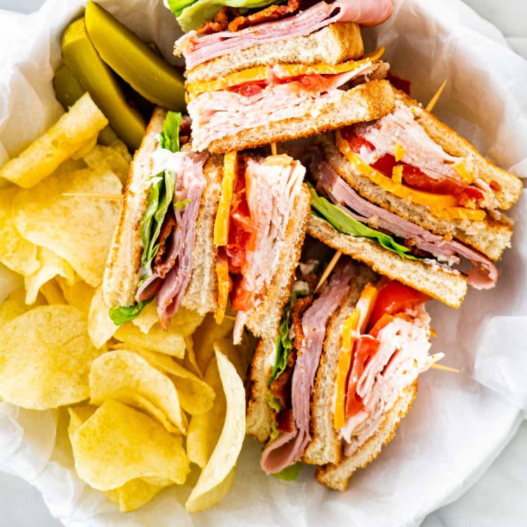 overhead of a club sandwich cut in quarters with a side of chips and pickles in a basket.