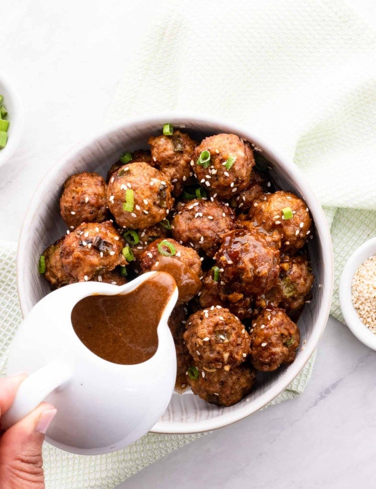 a hand holding a jug pouring an asian sauce over asian meatballs.