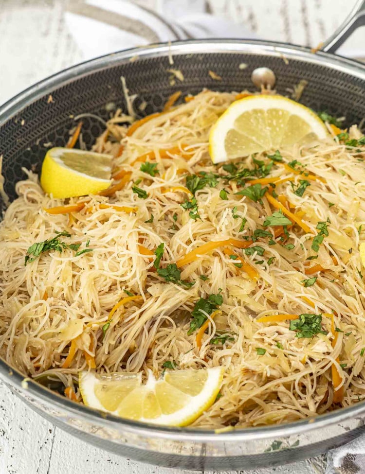 A skillet filled with Filipino pancit, thin rice noodles mixed with vegetables, garnished with lemon wedges and fresh herbs.