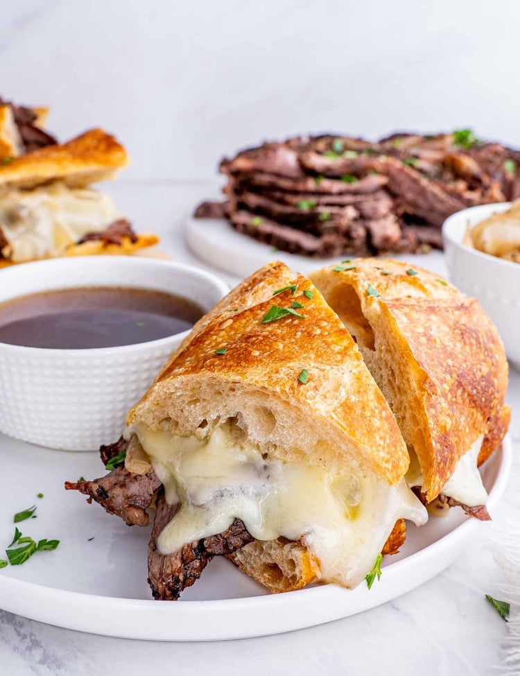 a french dip sandwich cu in half on a white plate next to a bowl with au jus.