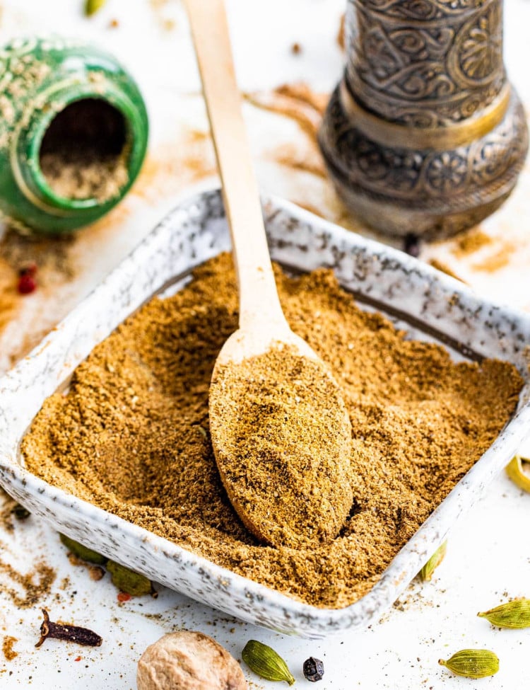 a square bowl filled with garam masala and a mini wooden spoon inside