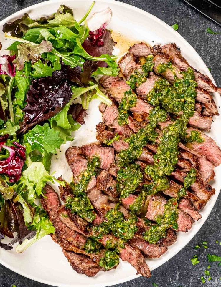 grilled ribeye sliced up drizzled with chimichurri sauce on a white plate along with a tossed green salad.