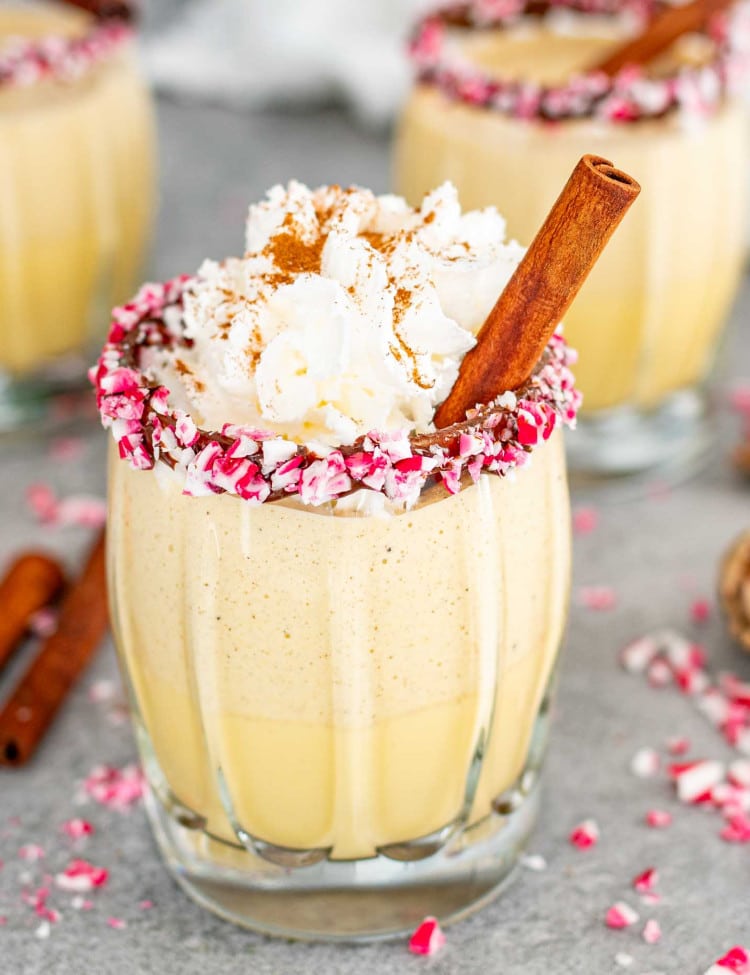 glasses of eggnog in glasses rimmed with chocolate and crushed candy cane pieces, garnished with cinnamon sticks.