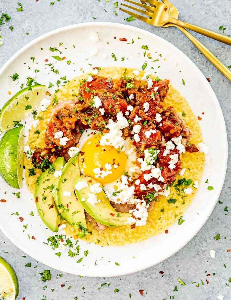 A vibrant plate of Huevos Rancheros featuring a sunny-side-up egg on a corn tortilla topped with refried beans, ranchero sauce, crumbled cheese, and avocado slices, garnished with fresh cilantro.