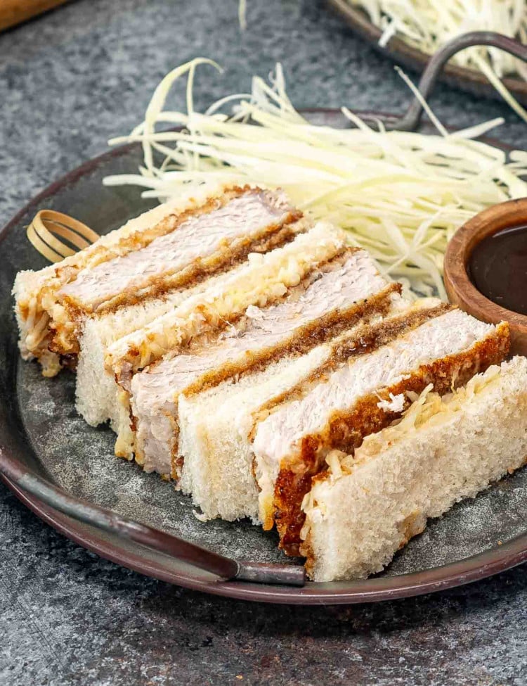 katsu sando on a metal plate with tonkatsu sauce in a bowl and cabbage.