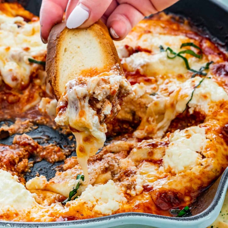 a hand dipping a piece of toasted bread into the lasagna dip