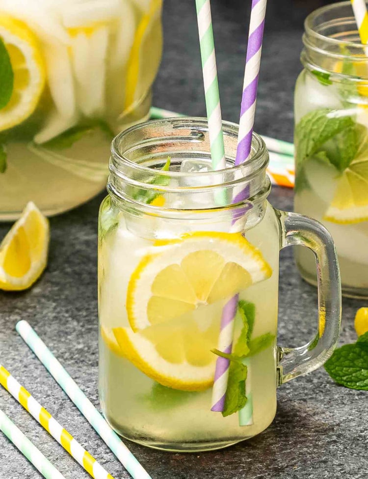 a jar full with lemonade, lemon slices, mint and a couple straws with a pitcher of lemonade in the background.