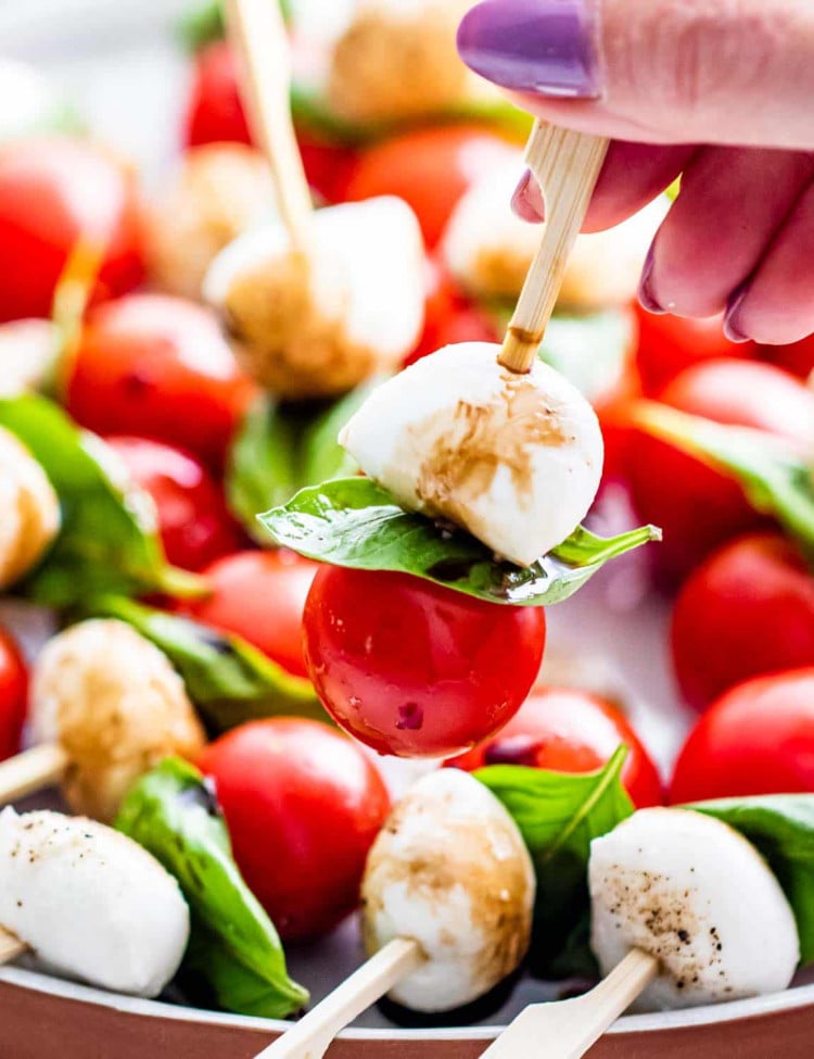 and hand holding a caprese salad bite on a pick over a plate full of them