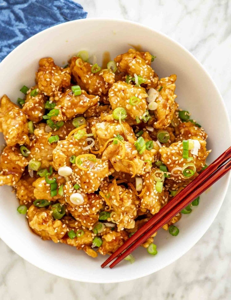 A bowl of crispy orange chicken topped with sesame seeds and finely chopped green onions. The chicken pieces are golden and glossy, coated in a rich orange sauce.