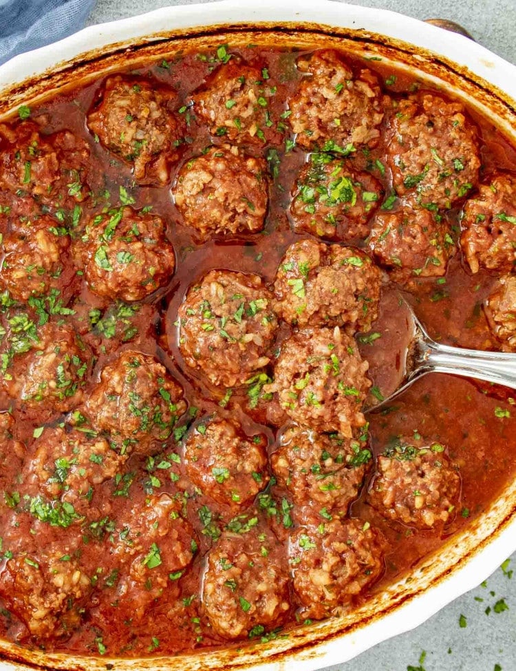 A dish of porcupine meatballs, cooked in a tomato-based sauce, garnished with fresh parsley, showcasing tender beef and rice-filled meatballs.