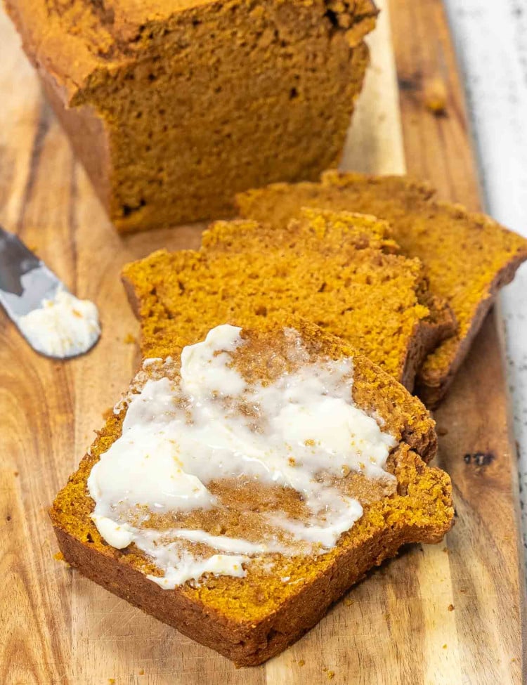 a few slices of pumpkin bread on a cutting board, one of them spread with butter.