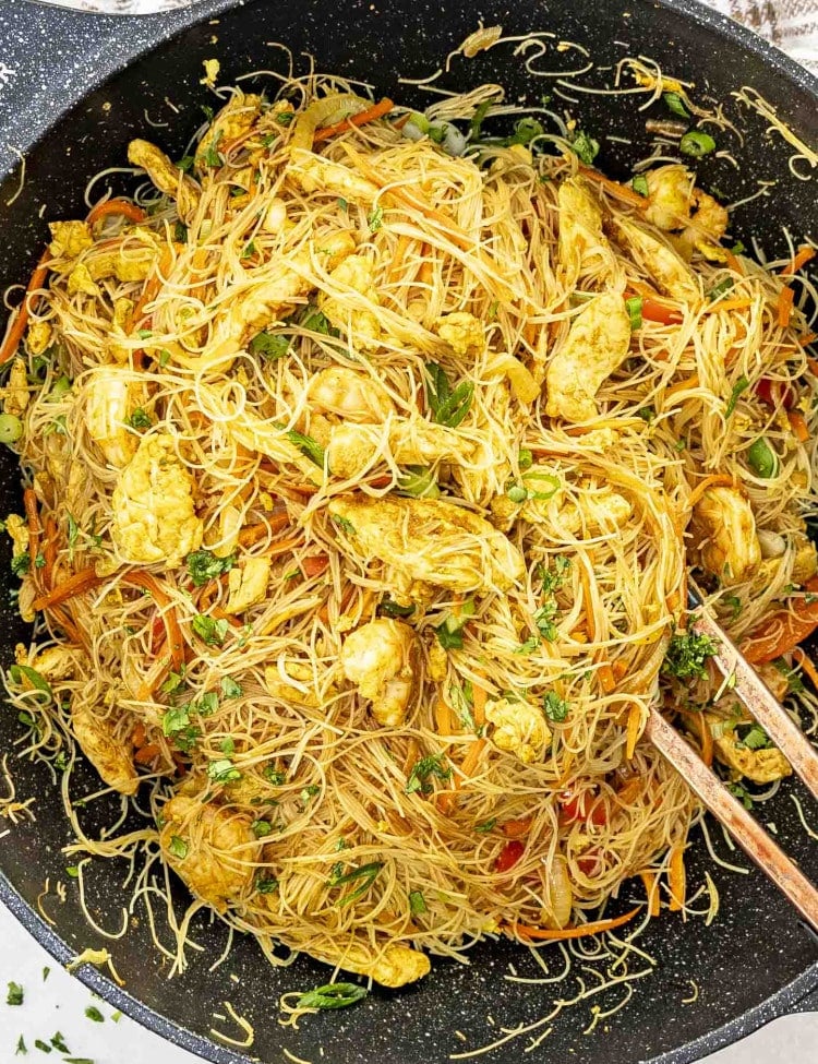 A vibrant dish of Singapore noodles served in a large black wok. The thin rice vermicelli noodles are mixed with chicken pieces, shrimp, and vegetables such as carrots, bell peppers, and green onions. The dish is seasoned with curry powder, giving it a yellowish hue, and garnished with fresh cilantro. Chopsticks are placed in the noodles, indicating it's ready to be served.