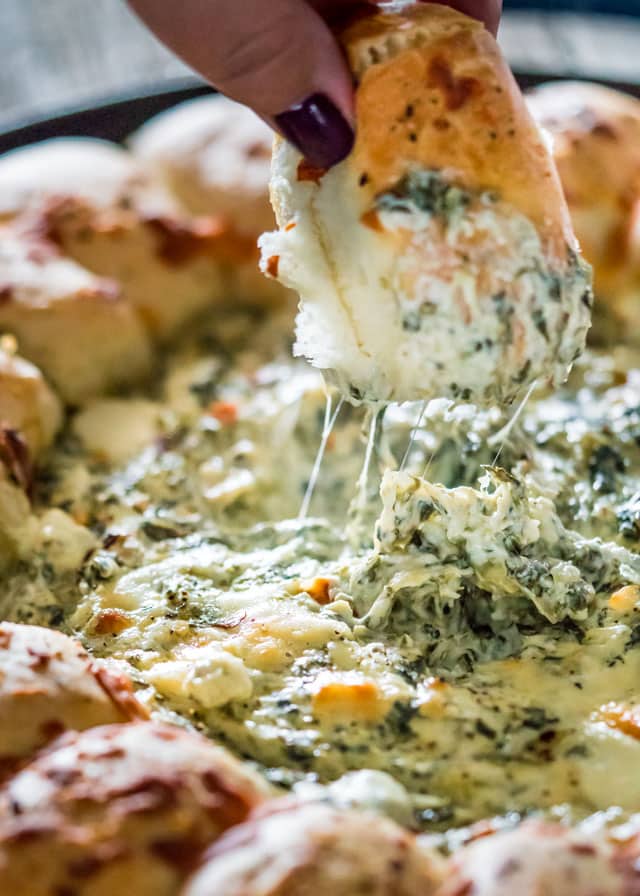 a hand picking up a bread roll from a pan of Spinach and Artichoke Dip