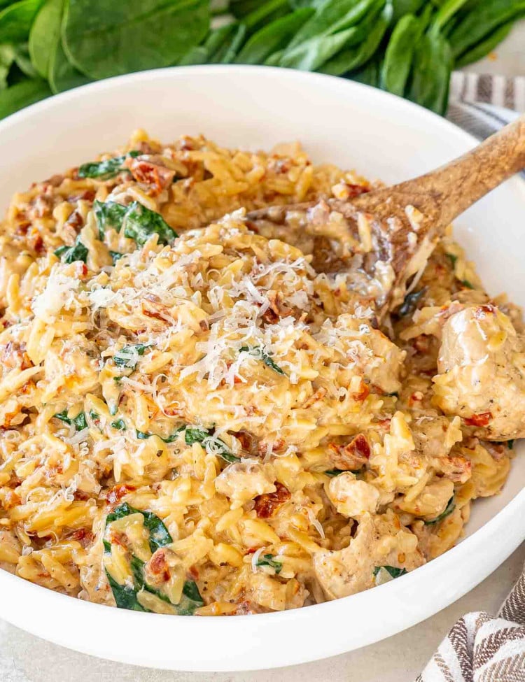 slow cooker creamy tuscan chicken orzo in a white serving bowl with a wooden spoon inside.