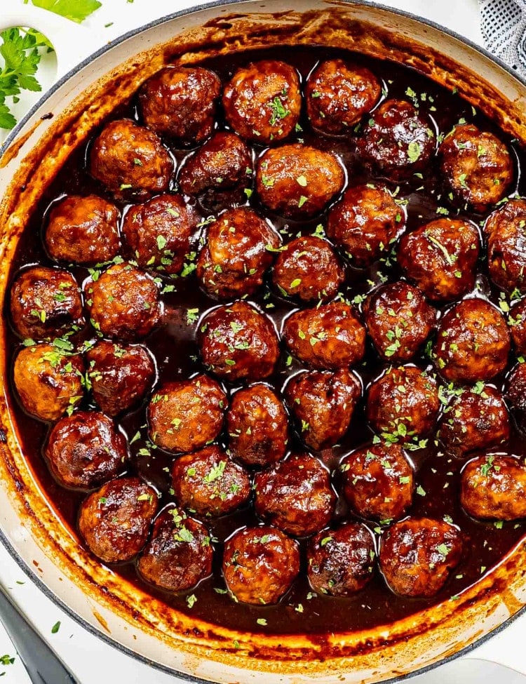 Savory stout meatballs, beautifully browned and simmering in a rich, smoky stout-infused BBQ sauce, served in a beige braiser, garnished with fresh parsley.