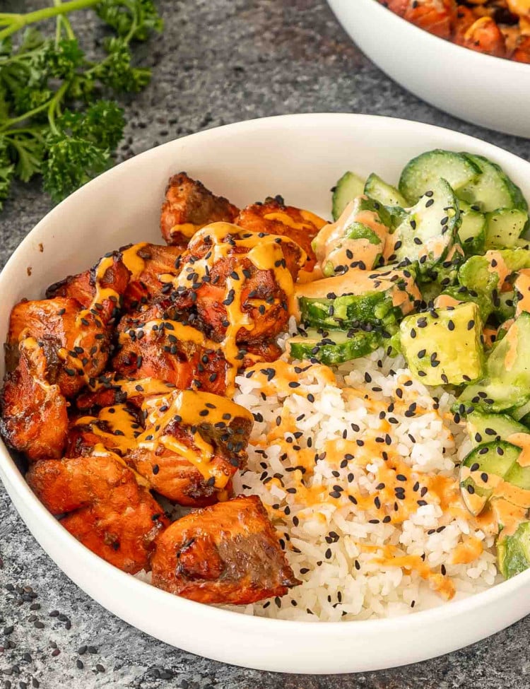 teriyaki salmon bites that were made in the air fryer, in a white bowl with steamed rice, cucumber avocado salad and a sriracha mayo.